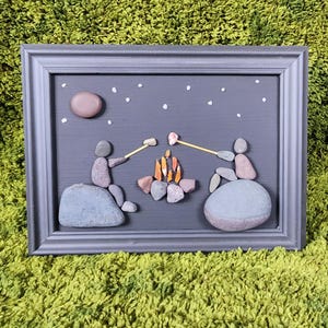 May include: Framed artwork depicting a campfire scene. Two figures made of stones sit around a campfire, roasting marshmallows. The background is a dark gray with a stone moon and white star accents. The frame is gray.