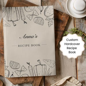 Peut inclure: Un livre de recettes personnalisé à couverture rigide avec le nom "Anna's" sur la couverture. La couverture beige présente des dessins au trait noirs de légumes et de pâtes. Les mots "RECIPE BOOK" sont en dessous du nom.