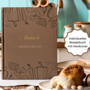 May include: A brown hardcover recipe book with the name "Anna's" in gold lettering. The cover features line drawings of vegetables and pasta. The text "Individuelles Rezeptbuch mit Hardcover" is in a white scalloped shape.