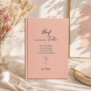 May include: A pink hardcover journal with the text "Brief an meine Tochter" in black script. The journal includes additional text and a floral illustration. The journal is displayed near a vase of dried flowers.