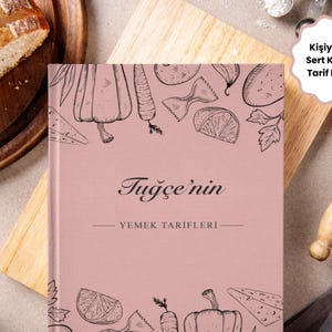 Könnte beinhalten: Ein rosa Kochbuch mit dem Titel "Tugce'nin" und den Worten "Yemek Tarifleri" darunter. Das Cover zeigt Strichzeichnungen von Gemüse und Pasta. Das Buch liegt auf einem Holzschneidebrett mit Brot und einer Teigrolle im Hintergrund.