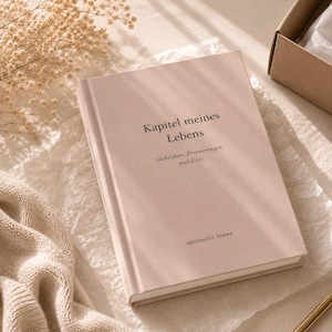 May include: A light pink hardcover journal with the German text "Kapitel meines Lebens" followed by "Gedanken, Erinnerungen und Ziele." and "optionaler Name" printed in black on the front cover. The book has a smooth, matte finish.