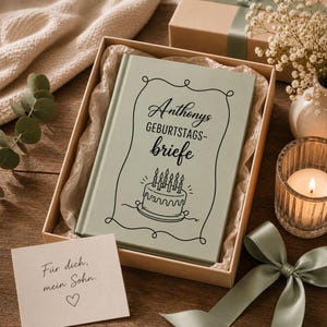 Puede incluir: Una caja de regalo que contiene un libro con el texto "Anthonys GEBURTSTAGS-briefe" y una ilustraci&oacute;n de pastel. Una peque&ntilde;a tarjeta dice "F&uuml;r dich, mein Sohn." La caja est&aacute; atada con una cinta verde, y una vela encendida y flores est&aacute;n cerca.