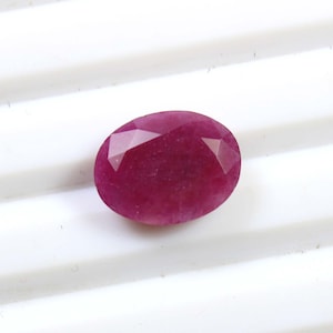 May include: A faceted, oval-shaped ruby gemstone. The gem is a deep, rich red color and has multiple facets that reflect light. The ruby is set against a white background.
