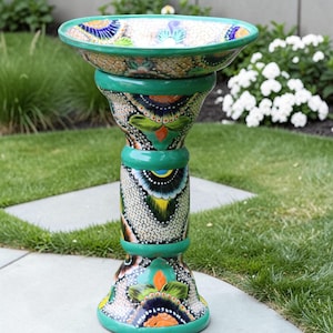 May include: A ceramic birdbath featuring a wide, shallow basin atop a cylindrical pedestal. The surface is decorated with intricate, colorful hand-painted floral and geometric patterns in shades of green, orange, yellow, blue, and white.