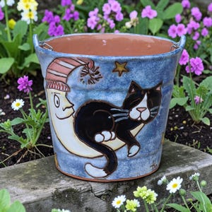 May include: A blue ceramic flower pot with a whimsical design. The pot features a black and white cat lounging on a crescent moon, a smiling moon wearing a nightcap, and stars. The pot has a brown interior and metal handles.