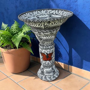 May include: A black and white bird bath with a decorative floral pattern and a red butterfly accent. The bird bath has a wide, shallow bowl and a pedestal base. A potted fern is to the left of the bird bath.