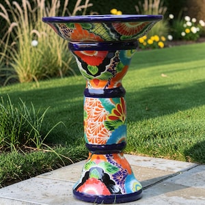 May include: A ceramic birdbath featuring a vibrant, multi-colored floral pattern with bold orange, blue, green, and yellow hand-painted details. The structure consists of a wide, shallow basin supported by a cylindrical pedestal and a flared base.