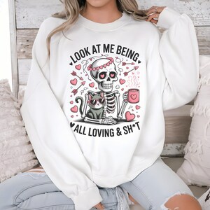 May include: White sweatshirt with a graphic featuring a skeleton with a floral crown, holding a mug, and a cat wearing sunglasses. The text reads "LOOK AT ME BEING ALL LOVING & SH*T" with hearts and arrows.