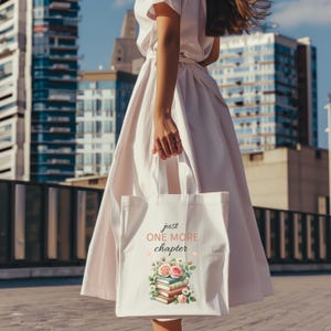 Puede incluir: Bolso tote de lona blanca con el texto "just ONE MORE chapter" y una ilustración de acuarela de libros y flores. El bolso lo sostiene una persona con un vestido blanco, con un horizonte urbano al fondo.