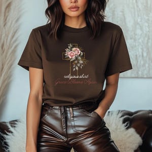 Puede incluir: Una camiseta marrón con un diseño de cruz dorada adornada con flores rosas y hojas verdes. El texto "religious shirt" y "Grace Blooms Again" está impreso debajo de la cruz.
