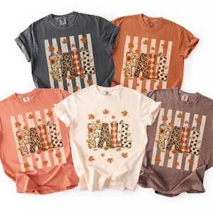 May include: Five short-sleeve t-shirts in charcoal, burnt orange, coral, cream, and brown. Each shirt features a central graphic with the word FALL in patterned letters, surrounded by vertical stripes and scattered autumn leaf illustrations.