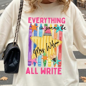 May include: An off-white t-shirt featuring a graphic of colorful crayons, a yellow star-shaped pencil, and small stars. The text reads "EVERYTHING is gonna be Mrs. Wilson ALL WRITE" in pink and black script and block lettering on the front.