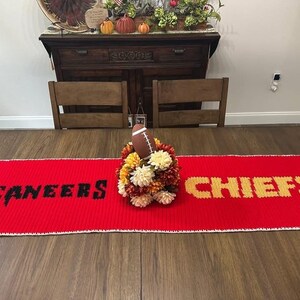 May include: A red table runner with the words "BUCCANEERS" and "CHIEFS" in black and gold, respectively. A football and floral arrangement sits in the center. The runner is on a wooden table.