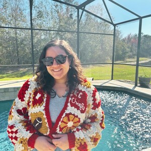 May include: A handmade crochet cardigan in red, white, and gold, featuring a football and the text "KC Chiefs". The cardigan has a V-neck and is made with a granny square pattern. The sleeves and trim are red.