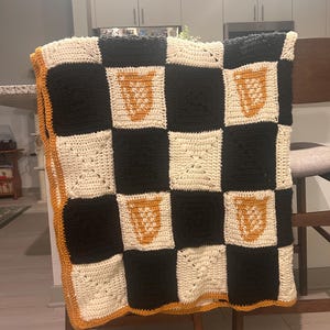 May include: A handmade crochet blanket with a checkerboard pattern in black, cream, and orange. The blanket features a repeating design of orange shapes on cream squares. The border is orange, and the blanket is draped over a wooden chair.