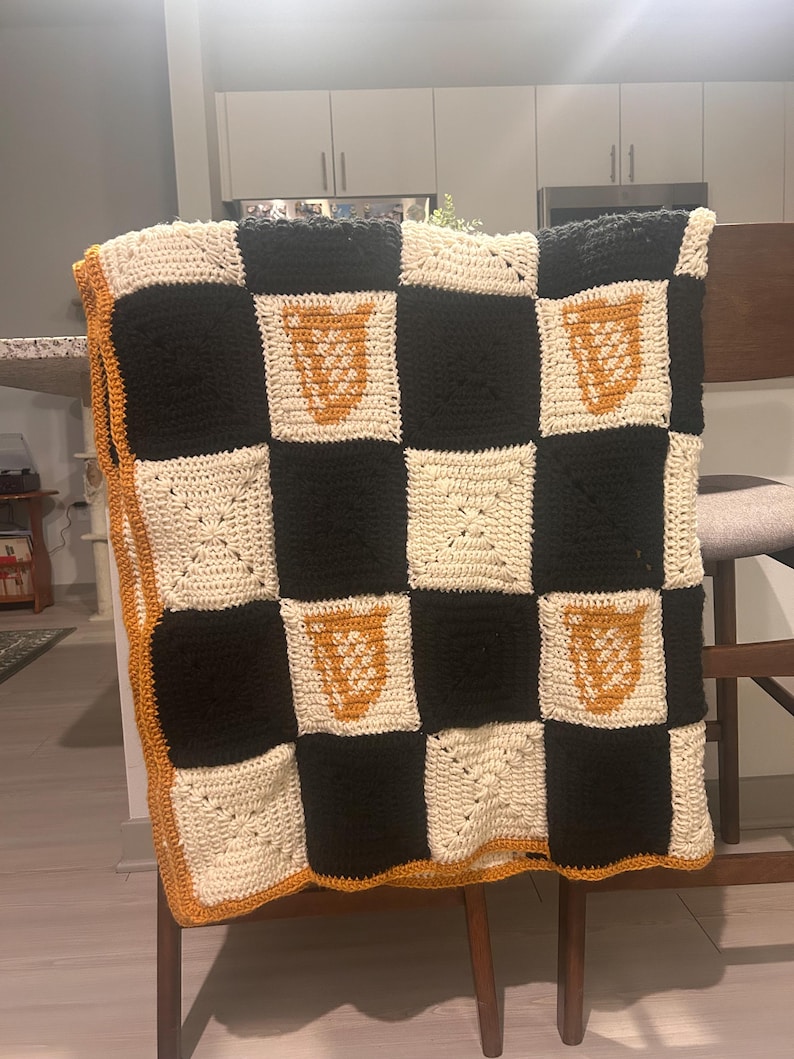 May include: A handmade crochet blanket with a checkerboard pattern in black, cream, and orange. The blanket features a repeating design of orange shapes on cream squares. The border is orange, and the blanket is draped over a wooden chair.