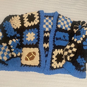 May include: A handmade crochet cardigan in a patchwork design. The color scheme includes black, blue, and beige squares, with a football and floral motifs. The cardigan has a blue trim and the text "MRS. L" on the front.