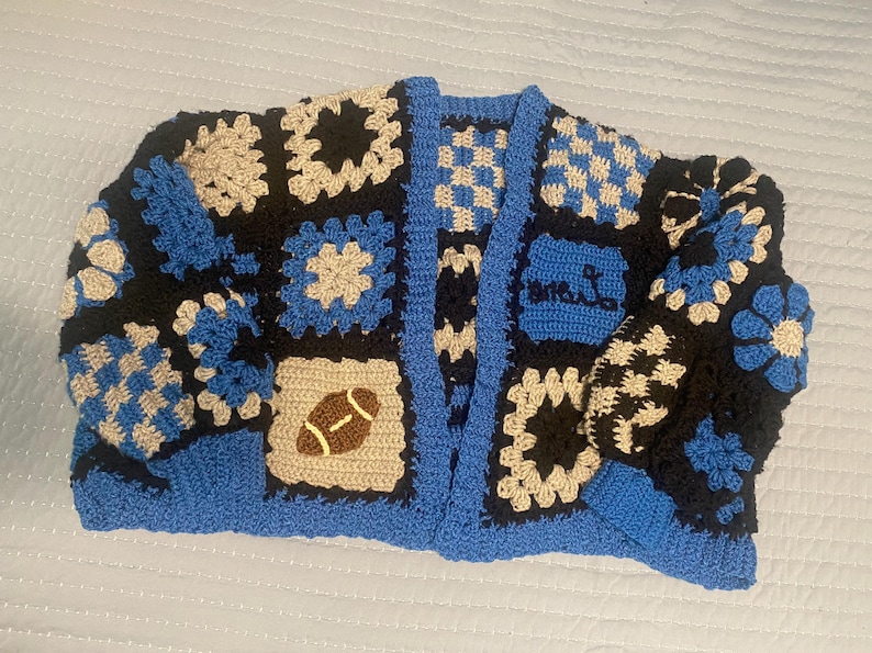 May include: A handmade crochet cardigan in a patchwork design. The color scheme includes black, blue, and beige squares, with a football and floral motifs. The cardigan has a blue trim and the text "MRS. L" on the front.