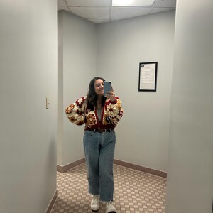May include: A person wearing a colorful crochet cardigan with a brown belt, blue jeans, and white sneakers. The cardigan features a patchwork design with red, cream, and brown squares. The jeans are a light wash with a raw hem.