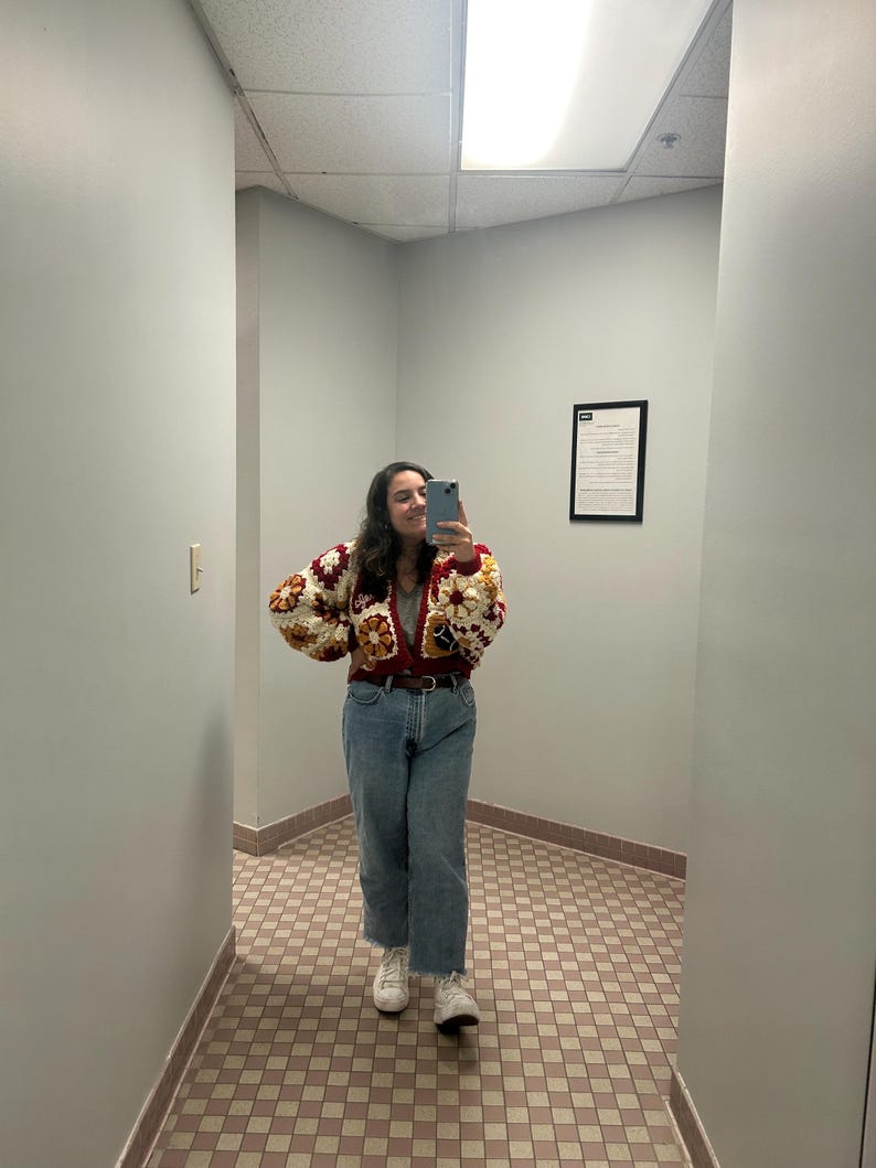 May include: A person wearing a colorful crochet cardigan with a brown belt, blue jeans, and white sneakers. The cardigan features a patchwork design with red, cream, and brown squares. The jeans are a light wash with a raw hem.