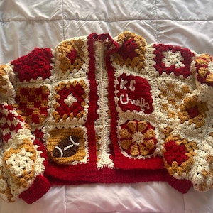 Könnte beinhalten: Handgefertigte Häkeljacke im Patchwork-Design. Die Jacke hat ein Farbschema aus Rot, Creme und Gold mit einem Football und dem Text "KC Chiefs". Die Jacke hat einen Reißverschluss und gerippte Bündchen.