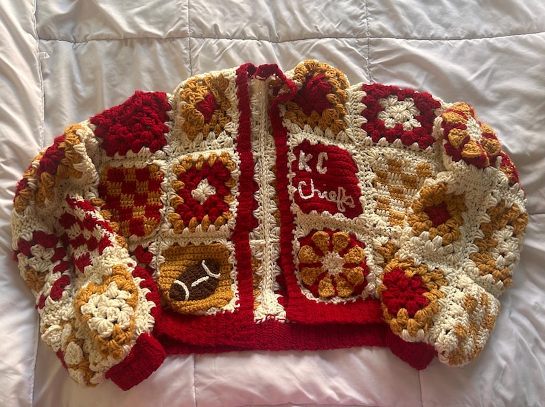 May include: A handmade crochet jacket in a patchwork design. The jacket features a red, cream, and gold color scheme with a football and "KC Chiefs" text. The jacket has a zipper closure and ribbed cuffs.