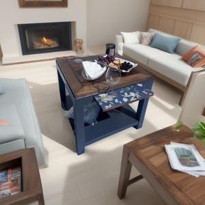 May include: A square coffee table with a dark blue base and a wooden top. The table has a lower shelf and a floral patterned pull-out tray. The table is set with a plate of food, glasses, and a napkin. A sofa and fireplace are in the background.