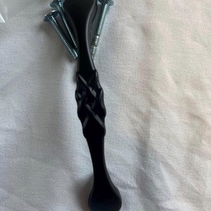 Puede incluir: Un tirador de armario negro con un diseño trenzado en el centro, acompañado de cuatro tornillos plateados. El tirador tiene un extremo en forma de lágrima y está sobre un fondo blanco.