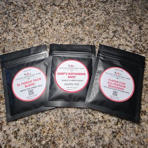 May include: Three black spice packets with white and red labels. The labels read "El Fuego Taco Blend", "Chef's Rotisserie Dust", and "Flavor Flex All-Purpose Seasoning". Each packet has the text "Leveling Up With Chef Jamie".