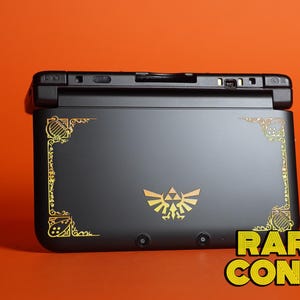 May include: A matte black handheld gaming console featuring a gold Triforce emblem in the center and ornate gold corner designs. The device has a hinge mechanism and two camera lenses on the bottom edge. The text RARETRO CONSOLES appears in yellow.