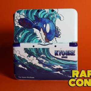 May include: A white handheld gaming console featuring a graphic of the blue Pokémon Kyogre emerging from stylized ocean waves. The text KYOGRE #382 is printed on the front, with The Hoenn Archives written in the bottom left corner.