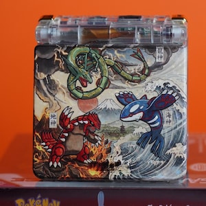 May include: A clear plastic handheld game console featuring a custom graphic on the lid. The artwork depicts three legendary creatures: a green serpentine dragon, a red bipedal monster, and a blue orca-like creature amidst waves, fire, and a mountain.