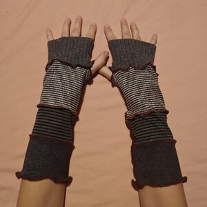 Könnte beinhalten: Ein Paar graue und gestreifte Fingerhandschuhe. Die Handschuhe bestehen aus verschiedenen Stoffabschnitten mit einem gerüschten Rand. Die Handschuhe sind grau, mit Abschnitten aus schwarz-weißen Streifen und einfarbigem Grau.