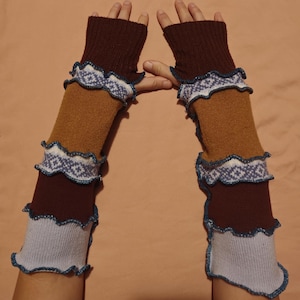 May include: A pair of knitted arm warmers in shades of brown, tan, and light blue. The arm warmers feature a ruffled edge with a white and blue geometric pattern. The cuffs are a darker brown color.