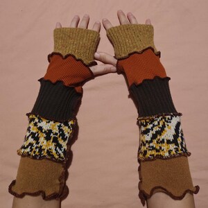 May include: Fingerless arm warmers in brown, orange, and black. The layered design includes a cuff with a yellow and black geometric pattern. The edges are finished with a ruffled trim. The arm warmers are designed to keep the arms warm.