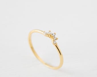 Puede incluir: Un delicado anillo de oro con una banda curva. El anillo presenta un grupo de pequeñas piedras preciosas transparentes dispuestas en un patrón floral o de estrella en la parte superior. El anillo está sobre un fondo blanco.