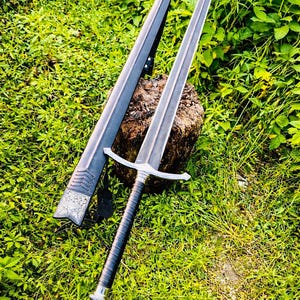 Pode incluir: Uma espada longa e prateada com uma empunhadura preta e uma bainha a condizer. A espada está apoiada num tronco de madeira rodeado por relva verde e folhagem. A bainha tem detalhes decorativos prateados.