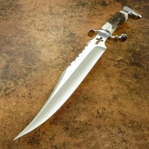 May include: A large, polished steel knife with a serrated edge and a cross emblem on the blade. The handle is made of a light-coloured material with dark brown accents. The knife has a silver guard and a curved end.