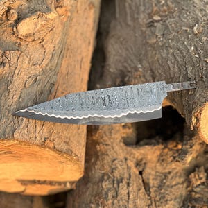 May include: A Damascus steel chef's knife with a patterned blade. The knife has a sharp, pointed tip and a full tang. The handle is not visible. The knife is set against a background of rough, brown tree trunks.