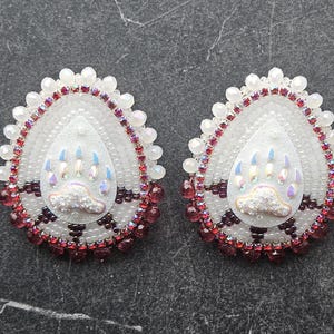 May include: A pair of teardrop-shaped beaded earrings featuring a central bear paw motif. The design incorporates white, iridescent, and deep red beads with a border of sparkling red crystals and white pearls, creating a detailed texture.