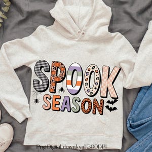 Peut inclure: Un sweat à capuche gris clair avec l'inscription "SPOOK SEASON" dans un design coloré et à motifs. Les lettres présentent des motifs tels que des carreaux, des imprimés léopard et des citrouilles. De petites toiles d'araignées, des araignées et des chauves-souris sont également incluses.
