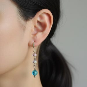 May include: A silver earring with a series of colourful beads in pink, clear, and blue. The earring is suspended from a silver hoop and is worn on a person's ear. The beads create a gradient effect.