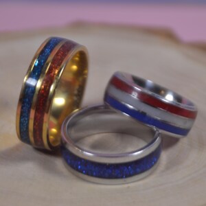 Könnte beinhalten: Drei Ringe mit farbigen Einlagen. Ein goldener Ring hat blaue und rote Einlagen. Zwei silberne Ringe haben blaue, rote und weiße Einlagen. Die Ringe werden auf einer Holzoberfläche präsentiert.