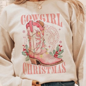 Sand beige sweatshirt with Western Christmas graphic design featuring a pink cowgirl boot wrapped in holiday lights, holly berries, rope lasso graphic, and text "Cowgirl Christmas"; festive Western holiday sweatshirt, country Christmas sweater, cowgirl aesthetic clothing, cozy winter pullover, Western Christmas gift, farmhouse holiday outfit, country girl Christmas apparel.
