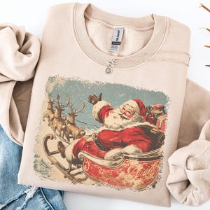 sand beige Christmas sweatshirt with vintage 60s Santa Claus riding sleigh and reindeer illustration; retro Santa Christmas sweater, classic holiday sweatshirt, cozy winter pullover, nostalgic Christmas design, unisex festive apparel, holiday gift idea, traditional Christmas aesthetic sweatshirt, matching family Christmas outfits.