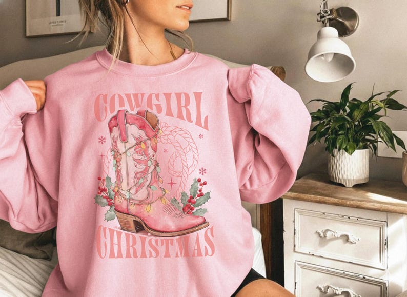 pink Sweatshirt with Western Christmas graphic design featuring a pink cowgirl boot wrapped in holiday lights, holly berries, rope lasso graphic, and text "Cowgirl Christmas"; festive Western holiday sweatshirt, country Christmas sweater, cowgirl aesthetic clothing, cozy winter pullover, Western Christmas gift, farmhouse holiday outfit, country girl Christmas apparel.