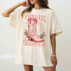 ivory comfort colors shirt with Western Christmas graphic design featuring a pink cowgirl boot wrapped in holiday lights, holly berries, rope lasso graphic, and text "Cowgirl Christmas"; festive Western holiday tee, country Christmas t-shirt, cowgirl aesthetic clothing, cozy winter sweatshirt, Western Christmas gift, farmhouse holiday outfit, country girl Christmas sweater.