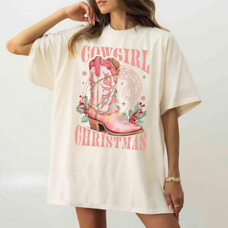 ivory comfort colors shirt with Western Christmas graphic design featuring a pink cowgirl boot wrapped in holiday lights, holly berries, rope lasso graphic, and text "Cowgirl Christmas"; festive Western holiday tee, country Christmas t-shirt, cowgirl aesthetic clothing, cozy winter sweatshirt, Western Christmas gift, farmhouse holiday outfit, country girl Christmas sweater.