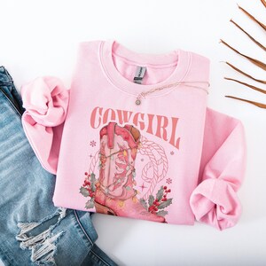 pink Sweatshirt with Western Christmas graphic design featuring a pink cowgirl boot wrapped in holiday lights, holly berries, rope lasso graphic, and text "Cowgirl Christmas"; festive Western holiday sweatshirt, country Christmas sweater, cowgirl aesthetic clothing, cozy winter pullover, Western Christmas gift, farmhouse holiday outfit, country girl Christmas apparel.